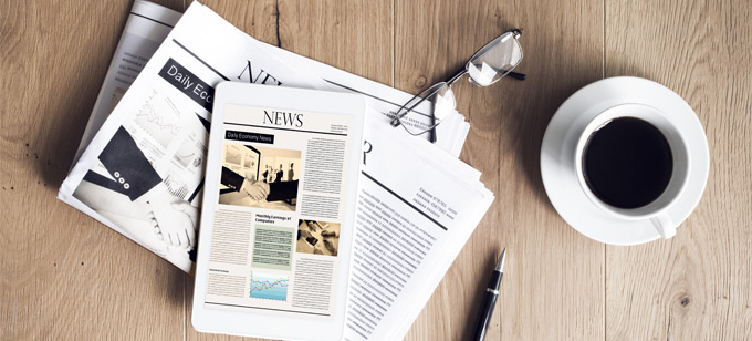 Newspaper and news website on tablet computer next to coffee and reading glasses on table. 